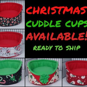 May include: Three Christmas-themed pet beds in the shape of a cuddle cup. The beds are red, green, and black with various Christmas patterns. The text "CHRISTMAS CUDDLE CUPS AVAILABLE! READY TO SHIP" is displayed above the beds.