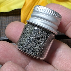 May include: A small glass vial with a silver lid, containing dark, granular material. The vial is held in a hand, with a yellow flower visible in the background. The contents appear to be a fine, dark powder or small particles.