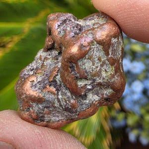 May include: A raw copper nugget with a metallic, reddish-brown surface featuring irregular, bumpy textures and patches of dark gray and green oxidation. The mineral specimen displays a natural, unrefined form with varied crevices and smooth protrusions.
