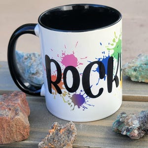 May include: White ceramic mug with a black handle and black rim. The mug has a colorful paint splatter design and the word "ROCK" in black lettering.