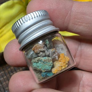 Puede incluir: Un pequeño frasco de vidrio con tapa plateada lleno de minerales coloridos. Los minerales incluyen tonos de turquesa, naranja, marrón y gris. El frasco está en la mano de una persona.