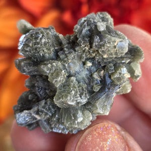 Op de afbeelding: Een close-up van een mineraal specimen met een complexe, gelaagde structuur. De steen heeft een gedempt groen- en grijstint, met kristallijne formaties. De achtergrond is wazig, met hints van oranje en rood.
