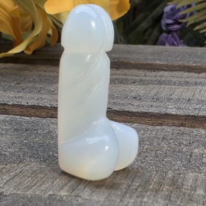 Könnte beinhalten: Eine glatte, cremefarbene Skulptur eines Phallus. Die Skulptur besteht aus einem durchscheinenden Material und hat einen abgerundeten Sockel und einen detaillierten Kopf. Das Objekt wird auf einer verwitterten Holzoberfläche ausgestellt.