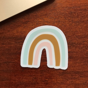 Könnte beinhalten: Ein Aufkleber mit einem Regenbogen in Pastellblau, Braun und Rosa. Der Regenbogen befindet sich auf einer Holzoberfläche.