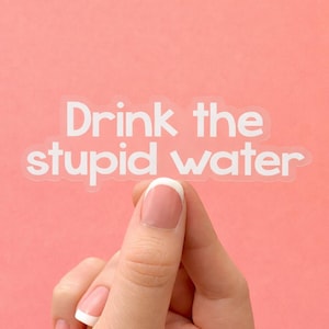 Puede incluir: Una pegatina de vinilo transparente troquelada que presenta el texto blanco "Drink the stupid water" en una fuente redondeada sin remates. La pegatina tiene un borde fino y transparente alrededor de las letras.