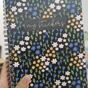 May include: A black spiral bound notebook with a floral pattern featuring white, yellow, blue, and green flowers. The notebook has a black label with the text "my stickers" written in white.