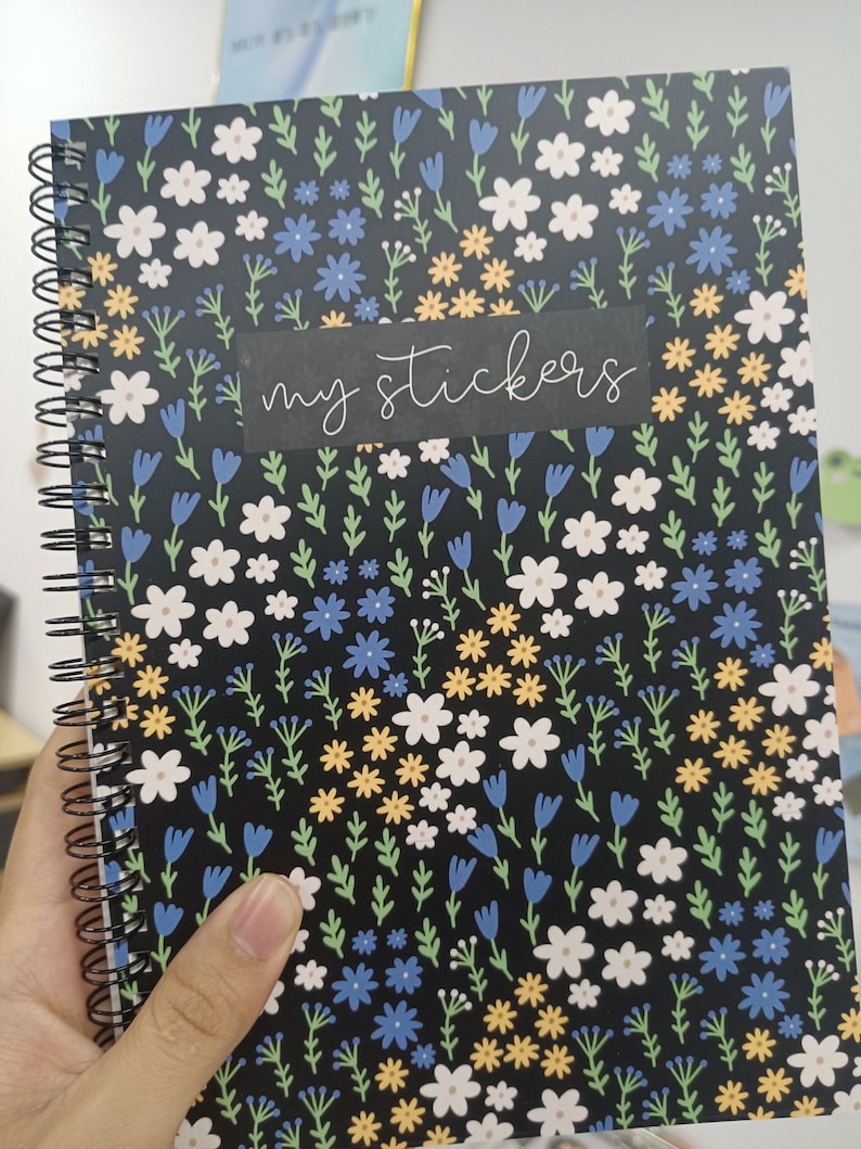 May include: A black spiral bound notebook with a floral pattern featuring white, yellow, blue, and green flowers. The notebook has a black label with the text "my stickers" written in white.