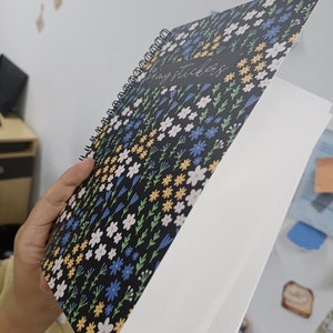 May include: A black spiral bound notebook with a floral pattern and the text "my stickers" on the cover. The notebook is open to a blank white page.