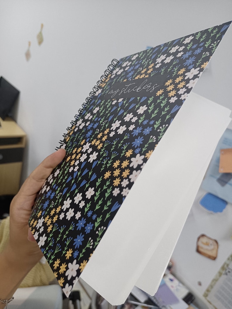 May include: A black spiral bound notebook with a floral pattern and the text "my stickers" on the cover. The notebook is open to a blank white page.