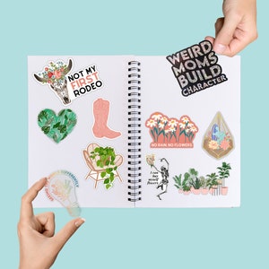 May include: A collection of colorful stickers featuring various designs, including a skull with flowers, a boot, a lightbulb, a chair with a plant, a skeleton holding flowers, a flower arrangement, and a terrarium. The stickers are arranged on a white notebook with a spiral binding. The text on the stickers includes "Not My First Rodeo", "Weird Moms Build Character", "Seeing the World Differently", "I can buy myself flowers", "No Rain. No Flowers".