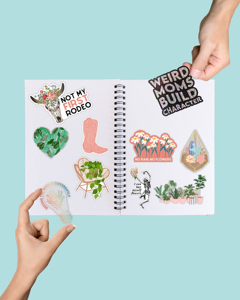 May include: A collection of colorful stickers featuring various designs, including a skull with flowers, a boot, a lightbulb, a chair with a plant, a skeleton holding flowers, a flower arrangement, and a terrarium. The stickers are arranged on a white notebook with a spiral binding. The text on the stickers includes "Not My First Rodeo", "Weird Moms Build Character", "Seeing the World Differently", "I can buy myself flowers", "No Rain. No Flowers".
