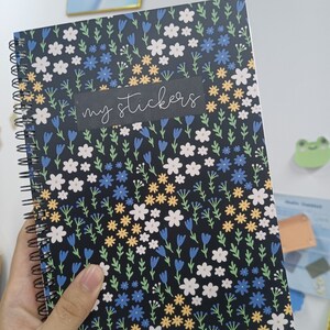 May include: A black spiral bound notebook with a floral pattern featuring white, blue, and yellow flowers. The notebook has the words "my stickers" written on a black label.