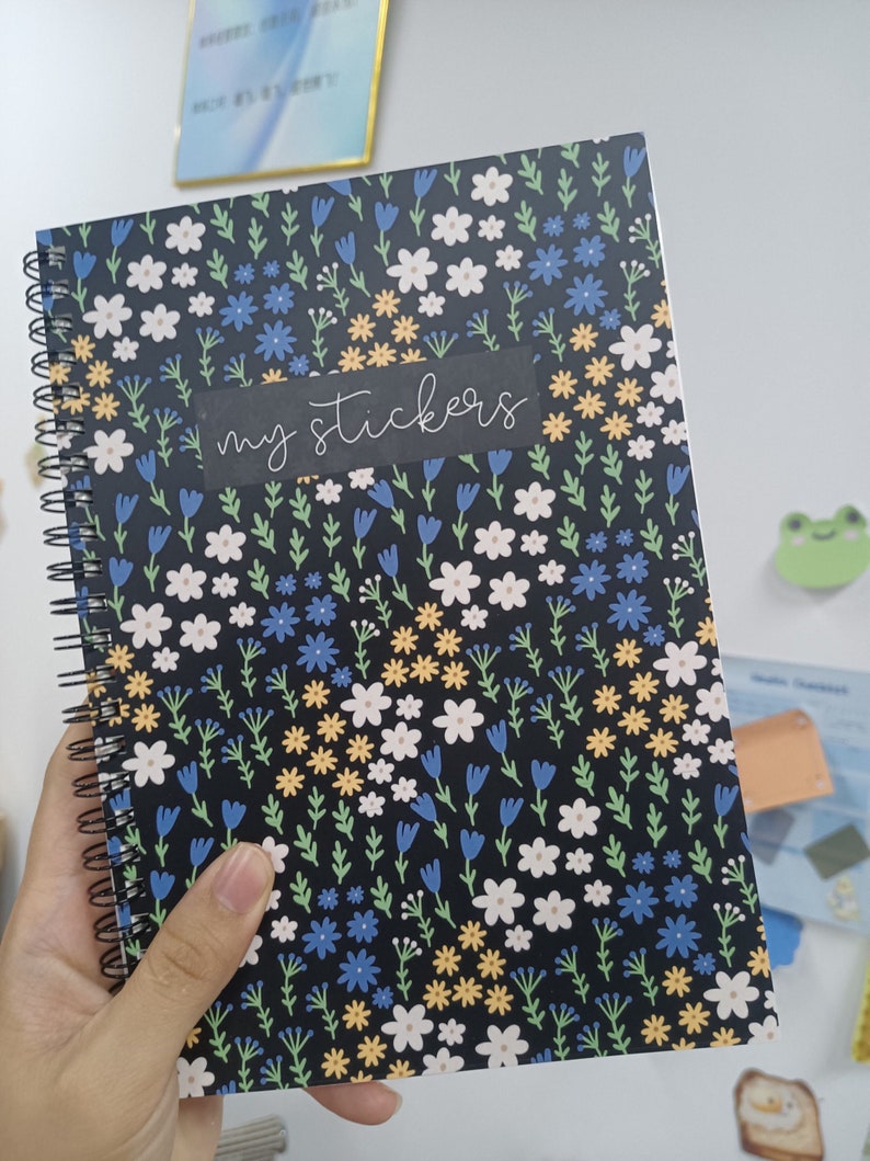 May include: A black spiral bound notebook with a floral pattern featuring white, blue, and yellow flowers. The notebook has the words "my stickers" written on a black label.