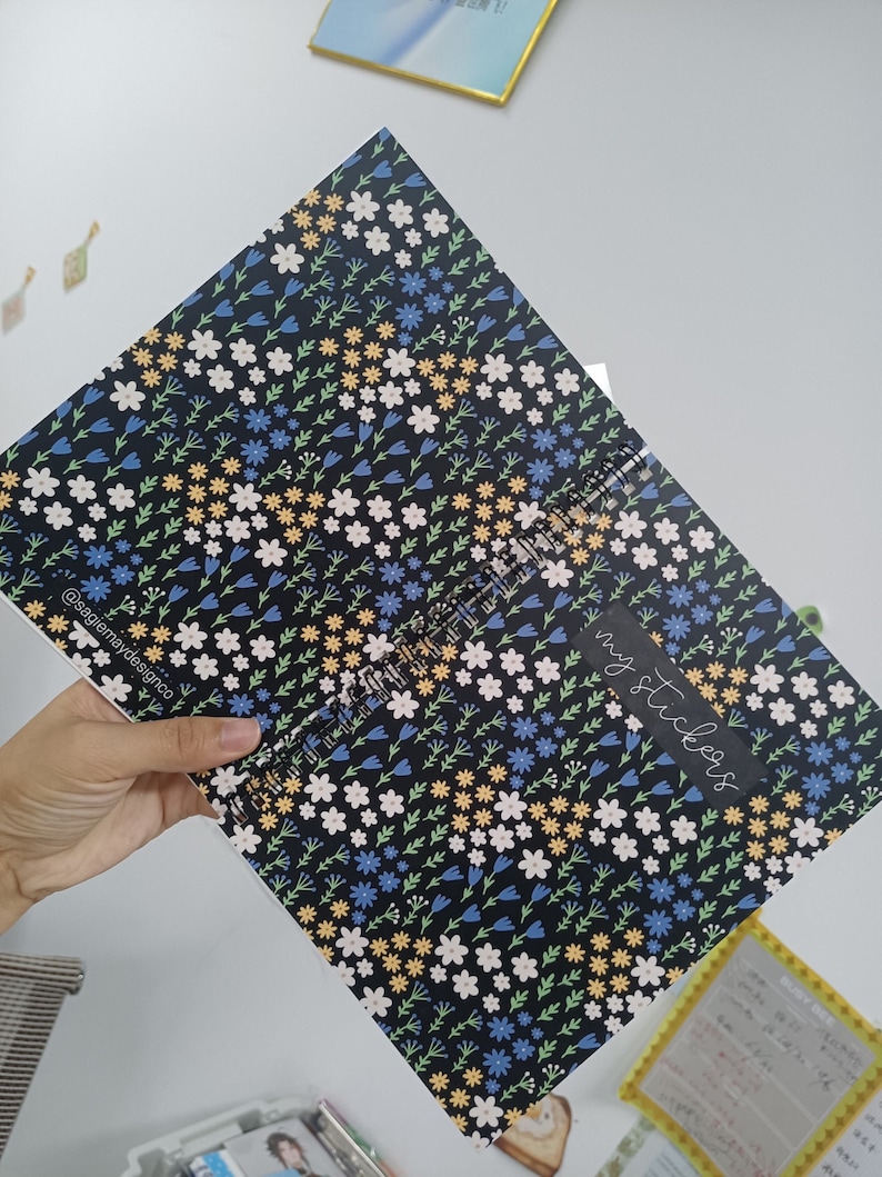 May include: A black spiral bound notebook with a floral pattern featuring white, blue, yellow, and green flowers. The text "my stickers" is written in white on a black rectangle on the cover.