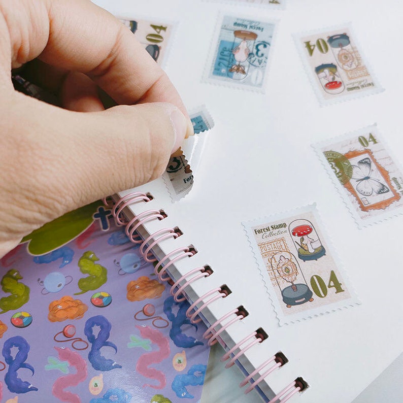 May include: A white spiral bound notebook with pink rings. The notebook has several colorful stickers on the pages. The stickers are of different shapes and sizes and feature images of butterflies, mushrooms, and other forest-themed designs. The stickers have the text "Forest Stamp Collection" and "Postage" printed on them.