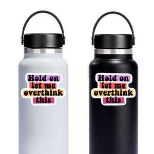 May include: Two stainless steel water bottles with black lids and handles. The bottles have stickers with the text "Hold on let me overthink this" in a retro font with colourful stripes.