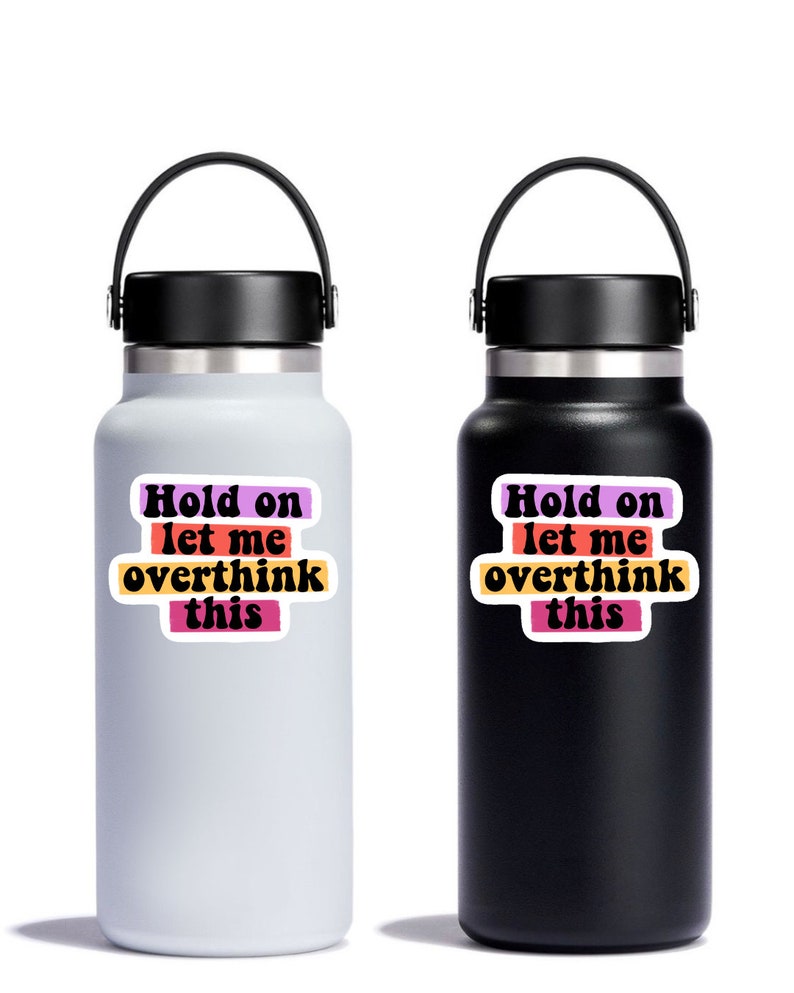 May include: Two stainless steel water bottles with black lids and handles. The bottles have stickers with the text "Hold on let me overthink this" in a retro font with colourful stripes.