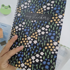 May include: A black spiral bound notebook with a floral pattern featuring white, yellow, blue, and green flowers. The notebook has a black label that says "my stickers".