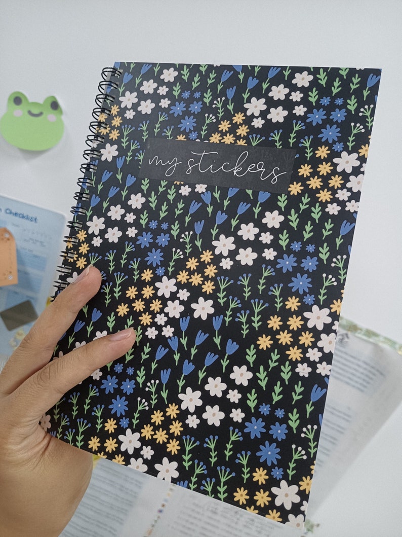May include: A black spiral bound notebook with a floral pattern featuring white, yellow, blue, and green flowers. The notebook has a black label that says "my stickers".