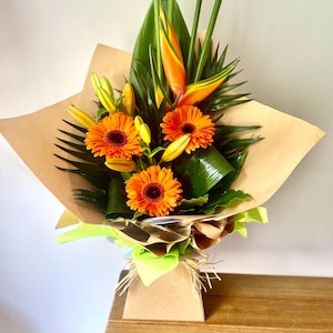 May include: A colourful bouquet featuring orange gerbera daisies, lilies, and exotic orange and yellow flowers, complemented by green foliage. The arrangement is wrapped in brown paper and presented in a decorative box, creating a visually appealing floral display.