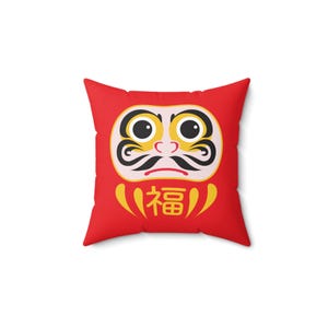 Pode incluir: Travesseiro quadrado vermelho com uma imagem de desenho animado de um amuleto de sorte japonês, um boneco Daruma, com rosto branco, sobrancelhas e bigode pretos, e grandes olhos pretos. O boneco usa uma túnica vermelha e tem a palavra japonesa "福" (que significa "boa sorte") escrita em sua barriga.