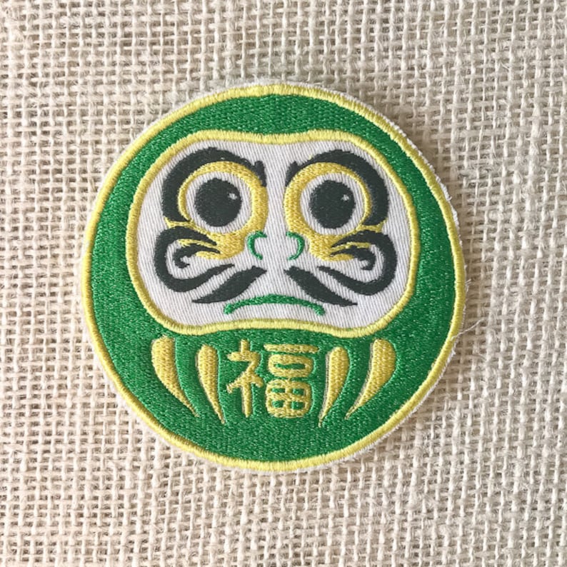 May include: Green and yellow embroidered patch of a Japanese Daruma doll with a white face and the word "福" (meaning "good fortune") written on the front.
