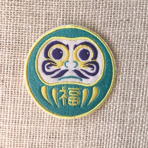 May include: A green and yellow embroidered patch of a traditional Japanese Daruma doll. The doll has a white face with purple eyes and a yellow mustache. The doll is holding a yellow scroll with the word "福" (meaning "good fortune") written on it.