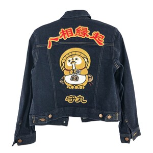 May include: A dark blue denim jacket with a yellow and red embroidered patch of a cartoon animal holding a small red bag. The text "~相 縁 起~" is embroidered in red on the jacket.