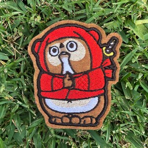 May include: Embroidered patch featuring a cartoon tanuki in a red hooded outfit. The tanuki has large, round eyes and is holding a small object. The patch has a brown border and is set against a background of green grass.