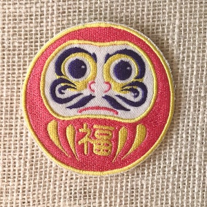 May include: A red and yellow embroidered daruma doll patch with a white face, black eyebrows, and purple eyes. The doll has a yellow "福" character on its belly.