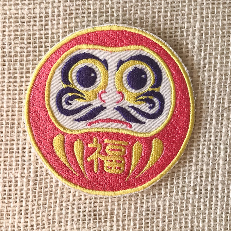 May include: A red and yellow embroidered daruma doll patch with a white face, black eyebrows, and purple eyes. The doll has a yellow "福" character on its belly.