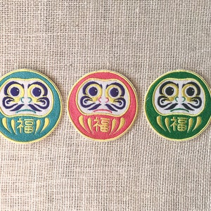 May include: Three round, embroidered patches featuring a traditional Japanese Daruma doll design. The Daruma dolls are depicted with a red, green, and blue background, and the word "福" (meaning "good fortune") is embroidered on each patch.