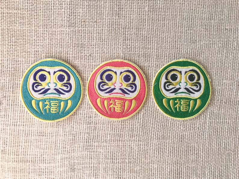 May include: Three round, embroidered patches featuring a traditional Japanese Daruma doll design. The Daruma dolls are depicted with a red, green, and blue background, and the word "福" (meaning "good fortune") is embroidered on each patch.