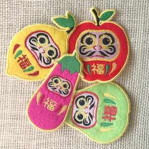Könnte beinhalten: Fünf bunte Filzpatches mit gestickten japanischen Daruma-Puppen. Jede Puppe hat eine andere Form, darunter eine Birne, eine Aubergine, ein Apfel und zwei runde Puppen. Die Puppen haben schwarze Augen, einen Schnurrbart und das Wort "福" (bedeutet "Glück") ist darauf gestickt.