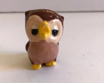 Puede incluir: Una pequeña figurita de búho de cerámica con plumas marrones y rosadas, ojos amarillos y un pico amarillo.