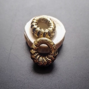 May include: Two gold-coloured metal rings with a textured, intricate design. The rings are stacked on top of each other on a white surface.