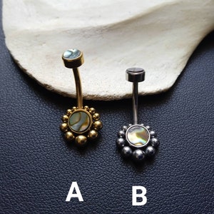Puede incluir: Dos anillos de ombligo con un diseño redondo de concha iridiscente. El anillo de la izquierda es dorado con una cadena de cuentas doradas. El anillo de la derecha es plateado con una cadena de cuentas plateadas. Los anillos están etiquetados como A y B.
