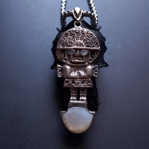 Op de afbeelding: Een zilverkleurige metalen hanger aan een ketting met een gestileerde figuur met ingewikkelde geëtste details, ingelegde witte schelpoogjes en een grote witte schelpvoet. De hanger hangt aan een zilverkleurige ketting met een schakelontwerp.