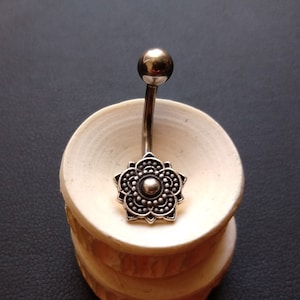 Op de afbeelding: Zilveren navelring met een decoratief bloemmotief. De ring ligt op een witte displaystandaard.