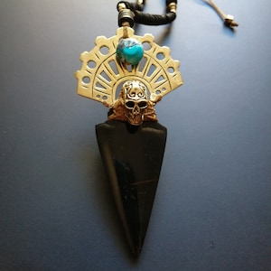 Peut inclure: Un collier artisanal avec un pendentif en forme de pointe de flèche noire, un crâne doré et une perle turquoise. Le collier est composé d'un cordon noir avec des perles dorées et d'un motif décoratif doré en forme d'éventail.