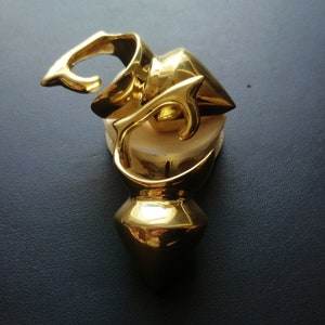 Può includere: Una scultura in metallo dorato con un design astratto. La scultura è realizzata in un unico pezzo di metallo e ha una finitura liscia e lucida.