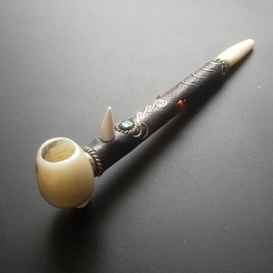 May include: A white bone pipe with a black and brown wooden stem. The stem is decorated with silver wire and red and green gemstones.