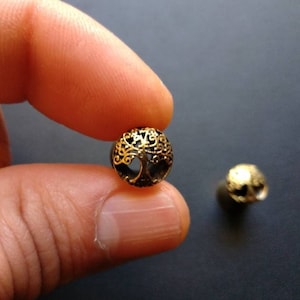 Könnte beinhalten: Zwei goldfarbene Metallperlen mit einem Lebensbaum-Design. Die Perlen sind rund und haben ein detailliertes, filigranes Design.