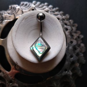 Puede incluir: Un piercing para el ombligo con una barra plateada y un colgante en forma de diamante de concha de abulón. La concha iridiscente refleja tonos de azul, verde y morado. La joya se exhibe sobre una superficie de cerámica blanca.