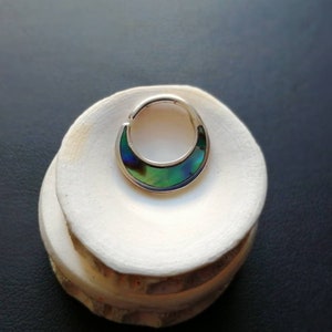 Peut inclure: Un anneau de septum argenté avec une incrustation en forme de croissant de coquille d'ormeau irisée. L'anneau est présenté sur une surface blanche et texturée, sur fond sombre. La coquille d'ormeau présente un mélange de teintes vertes, bleues et violettes.