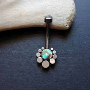 May include: A silver belly button ring with a central iridescent stone surrounded by small, circular silver accents. The jewelry is set against a dark background, with a white, textured object in the upper left corner.