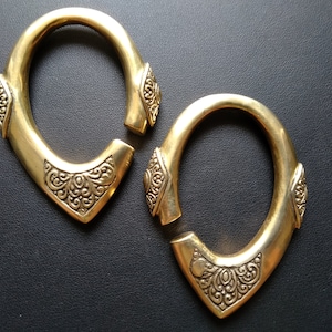 May include: Two gold-colored, oval-shaped earrings with a decorative, open design. Each earring features a detailed, ornate pattern at the bottom and on the sides. The earrings are displayed on a dark surface.