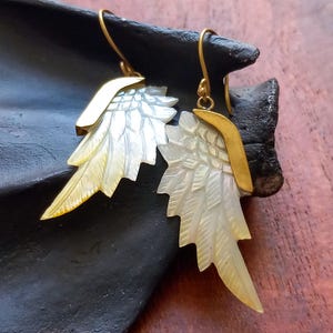 May include: A pair of earrings featuring a wing design. The earrings have a gold-colored metal top and a pearlescent, feather-like wing. The wings are white with yellow accents. The earrings are displayed on a dark surface.