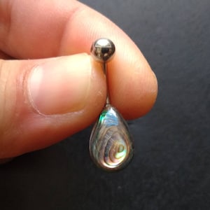 Puede incluir: Un piercing para el ombligo con un colgante en forma de lágrima de concha de abulón. El colgante tiene colores iridiscentes verdes, azules y dorados. La bola superior y el poste son de color plateado.