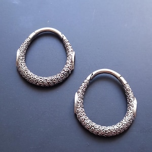 May include: A pair of silver-toned hoop earrings featuring a textured, scale-like pattern across the surface. Each hoop has a hinged closure mechanism and an elongated, teardrop-like shape with smooth, polished sections at the hinge and clasp points.